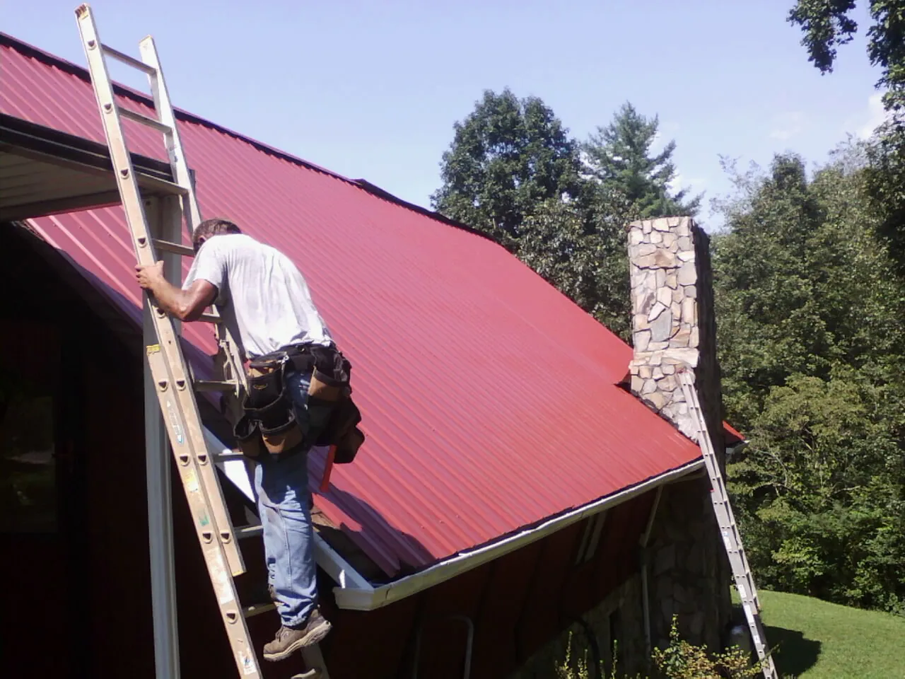 Roofer on a ladder beside a red metal roof and stone chimney — Soffit & Fascia Repair by Roofing Brothers in Tilden